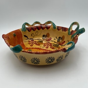 Puede incluir: Un bol de servir de cerámica amarillo en forma de pez con un diseño floral verde, rojo y naranja. El bol tiene dos asas y un borde festoneado.