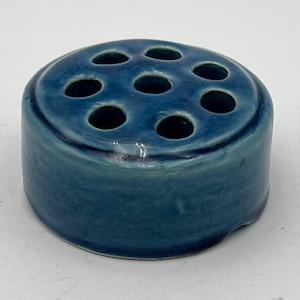 Könnte beinhalten: Ein blauer Keramik-Blumentopf mit acht Löchern zum Anordnen von Blumen. Der Topf ist rund und hat eine glatte, glasierte Oberfläche.