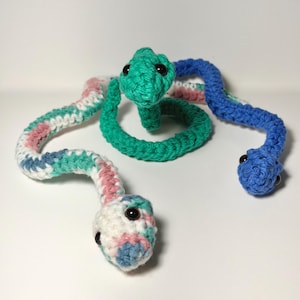 May include: Two crocheted snakes with black plastic eyes. One snake is solid teal and coiled, while the other is a long, winding snake featuring a variegated yarn pattern of white, pink, teal, and blue, ending in a solid blue tail section.