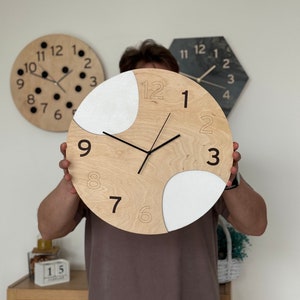 Pode incluir: Um relógio de madeira com detalhes brancos e números pretos. O relógio está sendo segurado por uma pessoa em frente a uma parede branca.