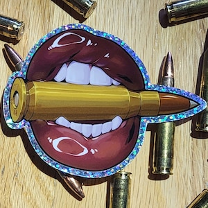 May include: A sticker of a red, glossy, open mouth with white teeth biting down on a gold bullet. The sticker has a holographic, iridescent border.