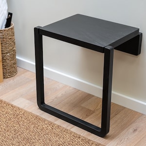 Puede incluir: Un banco negro montado en la pared con un asiento rectangular y un marco cuadrado abierto. El banco est&aacute; hecho de madera y metal. El banco mide 61 cm de largo, 30 cm de ancho y 46 cm de alto.