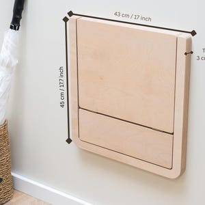 Puede incluir: Una mesa cambiador de pared de color claro hecha de madera. Mide 43 cm de ancho, 45 cm de alto y 3 cm de grosor. La mesa cambiador est&aacute; cerrada y montada en una pared blanca.