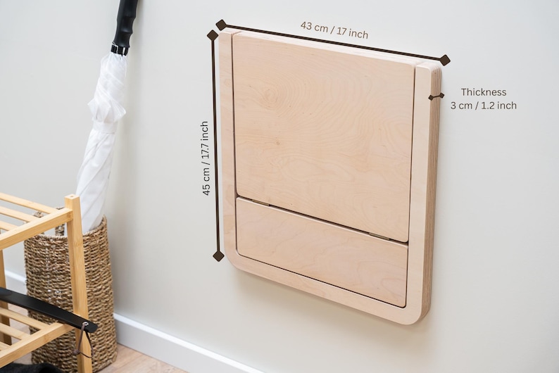 Puede incluir: Una mesa cambiador de pared de color claro hecha de madera. Mide 43 cm de ancho, 45 cm de alto y 3 cm de grosor. La mesa cambiador est&aacute; cerrada y montada en una pared blanca.