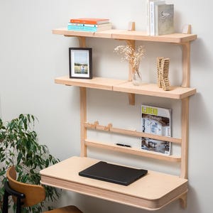 Pode incluir: Uma secretária de parede em madeira clara com prateleiras, uma pequena foto emoldurada e um laptop preto. A secretária tem uma prateleira embutida para revistas e uma caneta. Uma pequena cadeira de madeira está em frente à secretária.