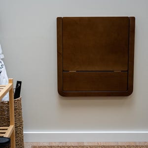 May include: A dark brown, wall-mounted folding table. The table is rectangular with rounded corners and a hinged design. The table is mounted on a light gray wall. A wooden shoe rack, a woven basket, and umbrellas are visible in the background.