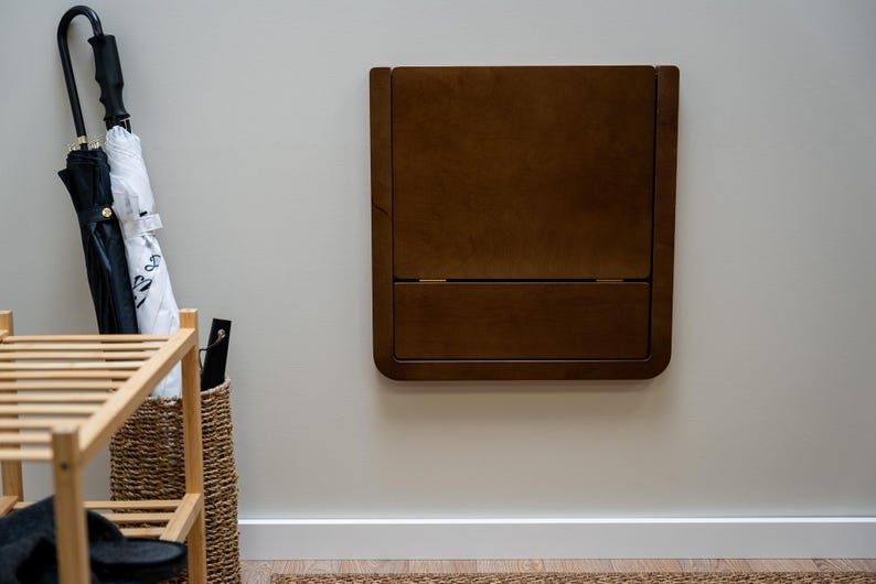 May include: A dark brown, wall-mounted folding table. The table is rectangular with rounded corners and a hinged design. The table is mounted on a light gray wall. A wooden shoe rack, a woven basket, and umbrellas are visible in the background.