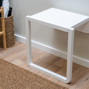 Op de afbeelding: Een witte, aan de muur gemonteerde klapbank met een vierkante zitting en een rechthoekig frame. De bank is gemaakt van hout en wit geschilderd. De bank is ontworpen om ruimte te besparen en functioneel te zijn.