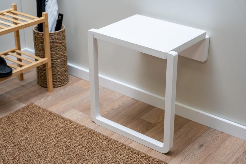 Puede incluir: Un banco plegable blanco, montado en la pared, con un asiento cuadrado y un marco rectangular. El banco est&aacute; hecho de madera y pintado de blanco. El banco est&aacute; dise&ntilde;ado para ahorrar espacio y ser funcional.