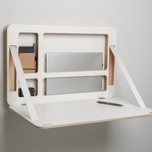 Könnte beinhalten: Weißer, an der Wand montierter Schreibtisch mit einer herunterklappbaren Oberfläche. Der Schreibtisch hat ein eingebautes Regal für einen Laptop und ein kleineres Regal für ein Notizbuch und einen Stift. Der Schreibtisch wird von zwei weißen Riemen gehalten.