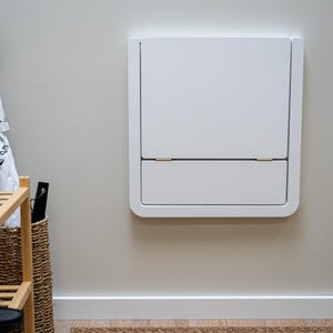 Puede incluir: Un armario blanco montado en la pared con un marco redondeado, con un compartimento superior e inferior. El armario est&aacute; montado en una pared de color beige claro. Se ve un paraguas blanco y negro y un zapatero de madera.