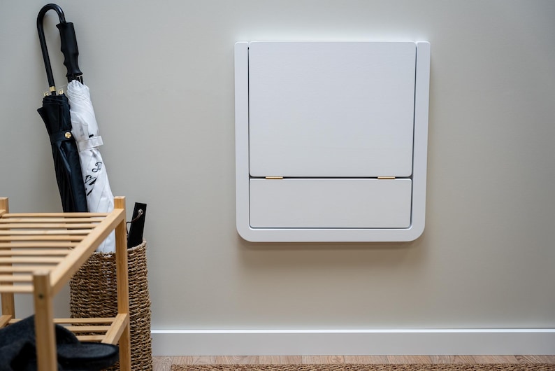 Puede incluir: Un armario blanco montado en la pared con un marco redondeado, con un compartimento superior e inferior. El armario est&aacute; montado en una pared de color beige claro. Se ve un paraguas blanco y negro y un zapatero de madera.