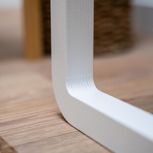 Puede incluir: Primer plano de una pata de mesa de madera pintada de blanco. La pata tiene un dise&ntilde;o curvo y est&aacute; colocada sobre un suelo de madera de color claro. La esquina de la pata es visible, mostrando la veta de la madera.