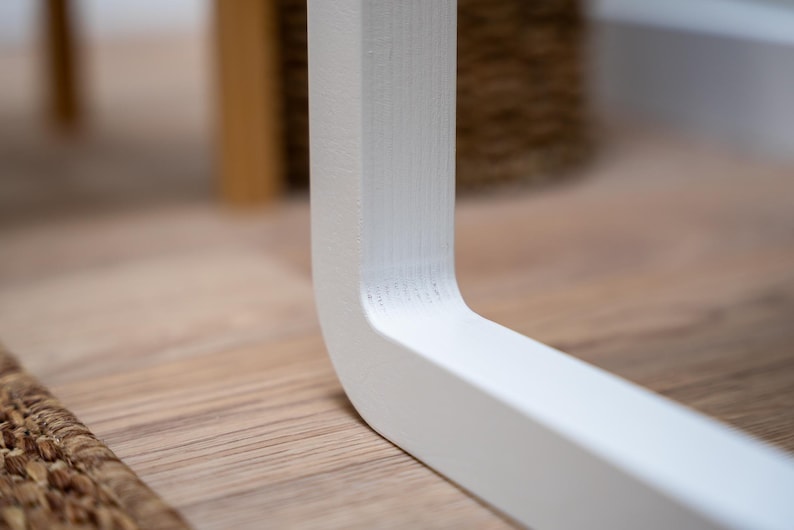 Puede incluir: Primer plano de una pata de mesa de madera pintada de blanco. La pata tiene un dise&ntilde;o curvo y est&aacute; colocada sobre un suelo de madera de color claro. La esquina de la pata es visible, mostrando la veta de la madera.