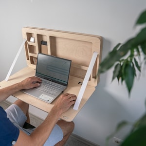 Könnte beinhalten: Ein heller Holz-Klapptisch an der Wand mit weißen Bändern. Ein Laptop ist auf dem Tisch geöffnet und eine Person benutzt ihn.