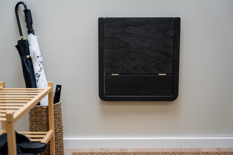 Puede incluir: Un mueble de pared de madera negra con una puerta con bisagras y un caj&oacute;n inferior. El mueble tiene un acabado de vetas de madera oscura y est&aacute; montado en una pared gris claro. Un zapatero, paraguas y una cesta de mimbre son visibles en el fondo.