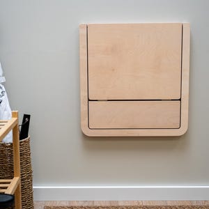 Puede incluir: Un escritorio plegable de color claro montado en la pared con esquinas redondeadas. El escritorio est&aacute; hecho de madera y tiene forma rectangular con una parte superior con bisagras y una secci&oacute;n inferior similar a un caj&oacute;n. El escritorio est&aacute; montado en una pared gris claro.