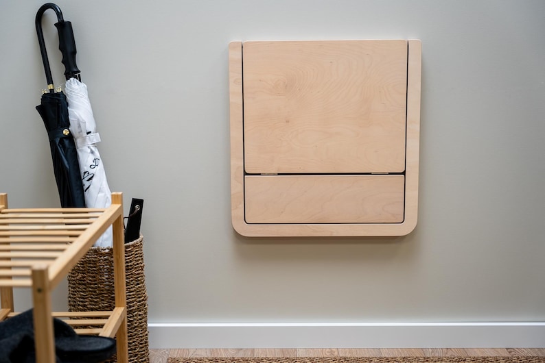 Puede incluir: Un escritorio plegable de color claro montado en la pared con esquinas redondeadas. El escritorio est&aacute; hecho de madera y tiene forma rectangular con una parte superior con bisagras y una secci&oacute;n inferior similar a un caj&oacute;n. El escritorio est&aacute; montado en una pared gris claro.