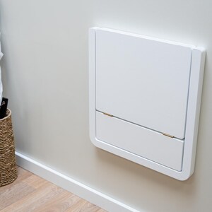 Puede incluir: Mesa plegable blanca, montada en la pared, en posici&oacute;n cerrada. Tiene forma rectangular con esquinas redondeadas y una peque&ntilde;a secci&oacute;n inferior con bisagras. Tambi&eacute;n se ve una cesta de mimbre con paraguas y un zapatero de madera.