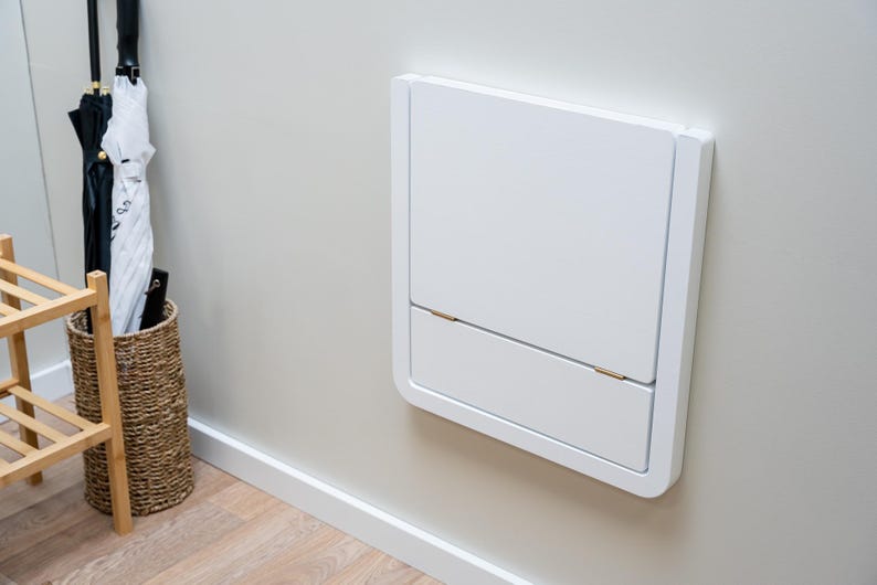 Puede incluir: Mesa plegable blanca, montada en la pared, en posici&oacute;n cerrada. Tiene forma rectangular con esquinas redondeadas y una peque&ntilde;a secci&oacute;n inferior con bisagras. Tambi&eacute;n se ve una cesta de mimbre con paraguas y un zapatero de madera.