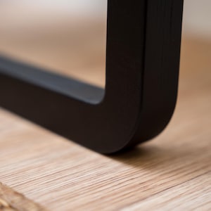 Puede incluir: Primer plano de una pata de mueble negra en &aacute;ngulo recto. La pata est&aacute; hecha de un material negro mate y descansa sobre una superficie de madera clara. La esquina de la pata es redondeada.