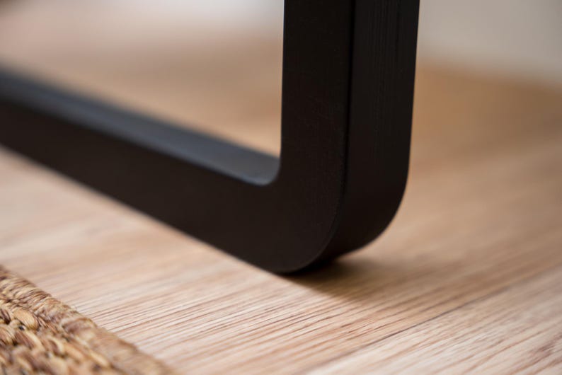 Puede incluir: Primer plano de una pata de mueble negra en &aacute;ngulo recto. La pata est&aacute; hecha de un material negro mate y descansa sobre una superficie de madera clara. La esquina de la pata es redondeada.