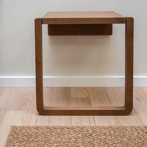 Puede incluir: Una mesa auxiliar de madera de forma cuadrada con un acabado marr&oacute;n c&aacute;lido. La mesa tiene un dise&ntilde;o minimalista con una parte superior rectangular y una base de marco abierto. La mesa mide aproximadamente 61 cm de alto y est&aacute; sobre un suelo de madera.