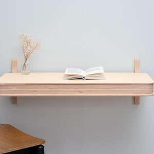Puede incluir: Un escritorio flotante de madera clara con bordes redondeados está montado en una pared gris claro. Un libro abierto y un pequeño jarrón de cristal con flores secas están sobre el escritorio. El escritorio está sostenido por dos soportes de madera.
