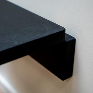 Puede incluir: Primer plano de una estanter&iacute;a flotante negra fijada a una pared blanca. La estanter&iacute;a tiene un dise&ntilde;o minimalista con una est&eacute;tica limpia y moderna. La estanter&iacute;a es de madera y tiene un acabado mate.