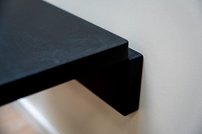 Puede incluir: Primer plano de una estanter&iacute;a flotante negra fijada a una pared blanca. La estanter&iacute;a tiene un dise&ntilde;o minimalista con una est&eacute;tica limpia y moderna. La estanter&iacute;a es de madera y tiene un acabado mate.