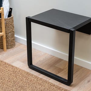 May include: A black, wall-mounted bench with a rectangular seat and a square, open frame. The bench is made of wood and metal. The bench is 24 inches long, 12 inches wide, and 18 inches high.