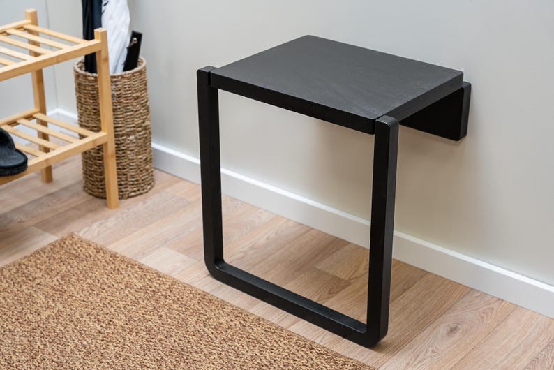 May include: A black, wall-mounted bench with a rectangular seat and a square, open frame. The bench is made of wood and metal. The bench is 24 inches long, 12 inches wide, and 18 inches high.