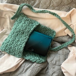 Puede incluir: Una bolsa de crochet verde azulado con una correa larga, que contiene una consola de juegos portátil Nintendo DS azul verdoso y negra.