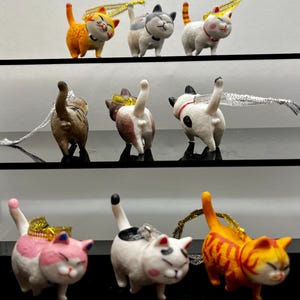 May include: A selection of cat ornaments in different colours and patterns. The ornaments are displayed on shelves, showing their front and rear views. Each cat has a small ribbon or string for hanging. The cats are orange, white, grey, and pink.