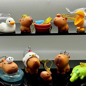 May include: A selection of small animal ornaments, featuring a white duck, brown capybaras, a pelican, and a green alligator. Each ornament is brightly coloured and has a silver or gold ribbon for hanging. These ornaments are suitable for festive decorations.