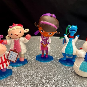May include: A collection of small character figurines, including a lamb, a snowman, and a dragon, all wearing medical coats. The figurines are on blue bases and have colorful details. The background is a sparkly silver surface.