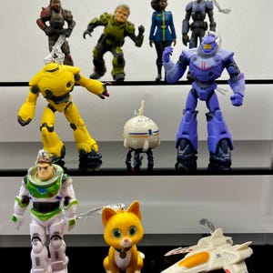 Puede incluir: Una colección de adornos de personajes, que incluyen un robot amarillo, un robot morado, una figura de Buzz Lightyear y un gato amarillo. Los adornos son de plástico y tienen cuerdas plateadas para colgar. Las figuras están en una estantería negra.