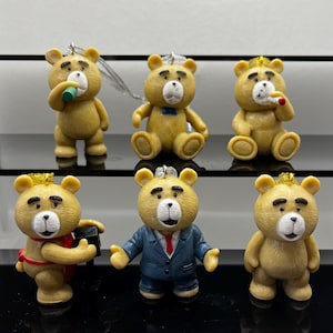 May include: Six small, tan-coloured bear figurines with various accessories. One bear holds a green cup, another a blue bow tie, and a third a cigarette. Other bears wear a crown, a suit, and a red scarf. Each has a silver string for hanging.