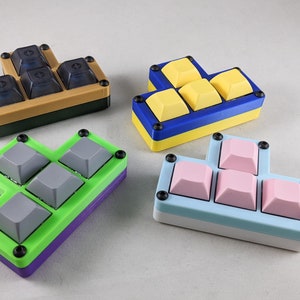 May include: Four different colored keyboard keycaps with different layouts. The keycaps are arranged in a T-shape, L-shape, and a staggered shape. The keycaps are made of a hard plastic and have a matte finish. The keycaps are in the colors of blue, yellow, green, and pink.