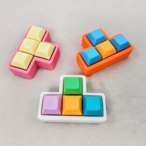 May include: Three colorful tile puzzles with different shapes and colors. The first puzzle is pink with yellow tiles, the second is orange with blue and orange tiles, and the third is white with green, orange, purple, and blue tiles.