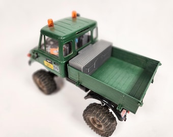 May include: A green toy truck with a flatbed and a gray toolbox in the back. The truck has orange lights on the cab and a "RACEL 06" sticker on the side.