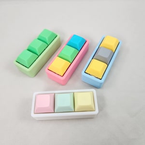 May include: Four sets of colorful, square-shaped keycaps in different colors, including green, blue, yellow, and gray. Each set has three keycaps and is arranged in a row. The keycaps are in a white, pink, and blue frame.