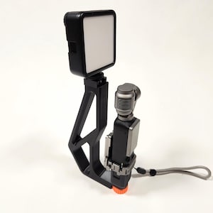 Op de afbeelding: Een zwart video-opname-apparaat met een witte LED-lamp, een microfoon en een grijze polsband. Het apparaat heeft een zwart frame met een oranje basis en is ontworpen voor video-opnamen.