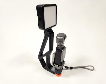 May include: A black video recording rig with a white LED light, a microphone, and a gray wrist strap. The rig has a black frame with an orange base and is designed for video recording.