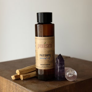 Puede incluir: Una botella de vidrio marr&oacute;n con una tapa negra y una etiqueta que dice "purism Palo Santo sacred oil massage & body oil". La botella est&aacute; sobre una superficie de madera con tres palitos de Palo Santo y dos cristales.