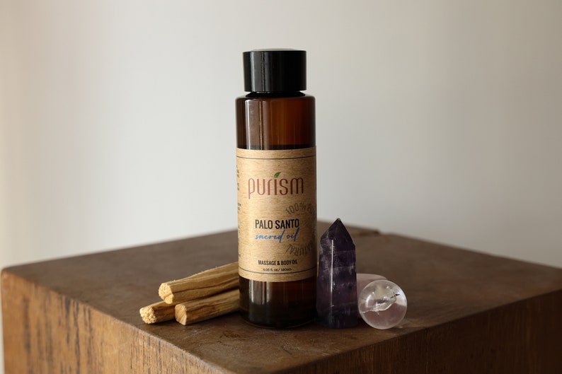 Puede incluir: Una botella de vidrio marr&oacute;n con una tapa negra y una etiqueta que dice "purism Palo Santo sacred oil massage & body oil". La botella est&aacute; sobre una superficie de madera con tres palitos de Palo Santo y dos cristales.