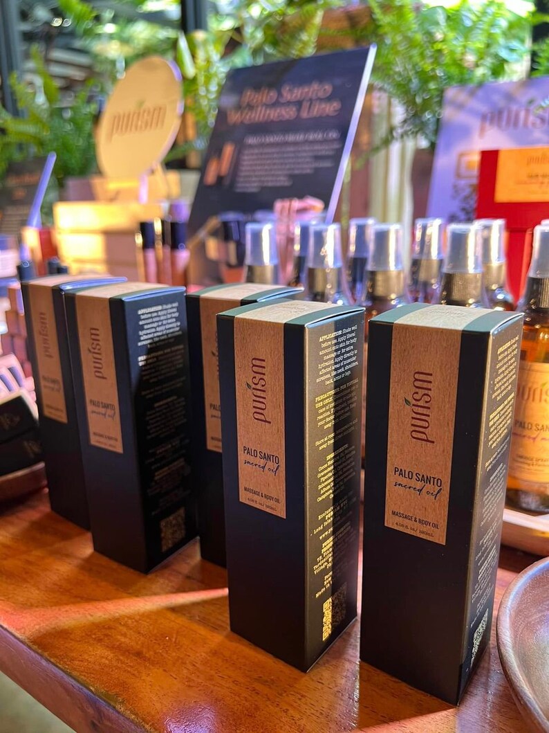 Puede incluir: Cuatro botellas negras de aceite sagrado de Palo Santo para masaje y cuerpo, con letras doradas y un c&oacute;digo QR en la etiqueta. Las botellas est&aacute;n dispuestas en fila sobre una superficie de madera. Las botellas est&aacute;n etiquetadas con "purism" y "Palo Santo sacred oil".
