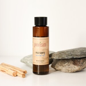 Puede incluir: Una botella de vidrio marr&oacute;n con una tapa negra y una etiqueta que dice "purism 100% pure natural PALO SANTO sacred oil MASSAGE & BODY OIL 10 ml (0.34 fl oz)". La botella est&aacute; sentada sobre una pila de rocas grises.