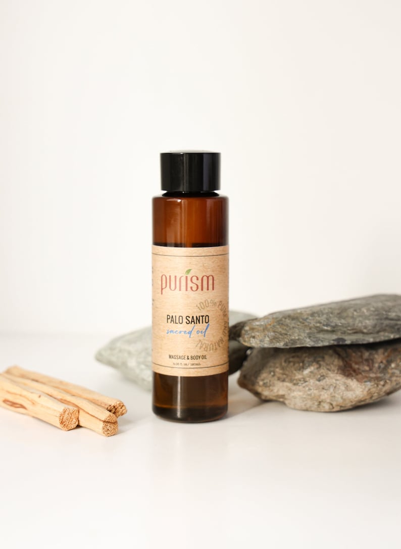Puede incluir: Una botella de vidrio marr&oacute;n con una tapa negra y una etiqueta que dice "purism 100% pure natural PALO SANTO sacred oil MASSAGE & BODY OIL 10 ml (0.34 fl oz)". La botella est&aacute; sentada sobre una pila de rocas grises.