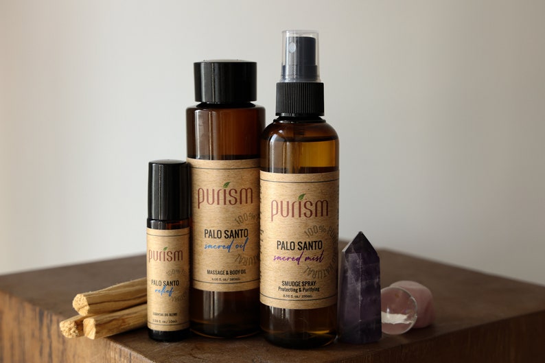 Puede incluir: Tres botellas de vidrio marr&oacute;n con etiquetas que dicen "purism" y "Palo Santo" en varios tama&ntilde;os. Las botellas contienen aceite sagrado de Palo Santo, bruma sagrada y aceite de rodillo. Las botellas est&aacute;n dispuestas sobre una superficie de madera con palitos de incienso de Palo Santo y un cristal morado.